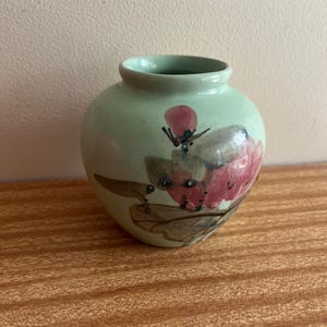 Könnte beinhalten: Eine kleine, hellgrüne Keramikvase mit einem Blumendesign. Die Vase hat einen runden Körper und eine schmale Öffnung. Das Blumendesign ist in Rosa-, Braun- und Schwarztönen bemalt.