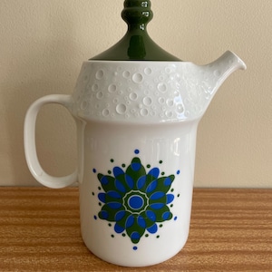 May include: White ceramic teapot with a green lid and handle. The teapot has a blue and green floral design. The top has a textured, dotted pattern. The spout is white and angled. A decorative item for the kitchen.