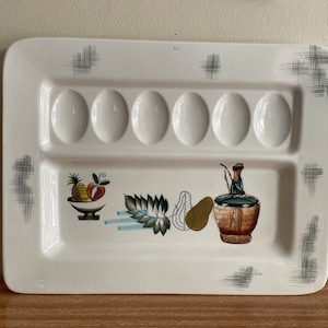 May include: Rectangular white ceramic serving platter with seven egg indentations and fruit illustrations. The platter features a pineapple, apple, pear, and a bottle with a woven basket design. Gray crosshatch accents adorn the corners.