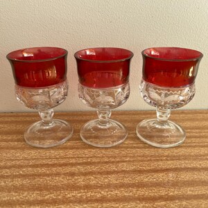 Può includere: Tre calici vintage in vetro rosso rubino e trasparente. La parte superiore di ogni calice è di un rosso intenso, mentre la parte inferiore è trasparente con un motivo decorativo. I calici sono esposti su una superficie di legno.