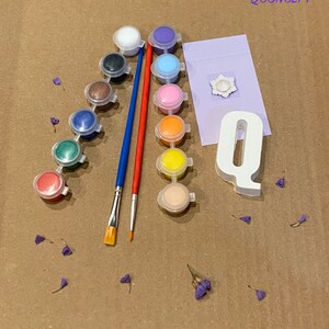 May include: A paint-by-numbers kit featuring a white letter Q, a small white flower, and 12 paint pots in various colours. The kit also includes two paintbrushes, one red and one blue.