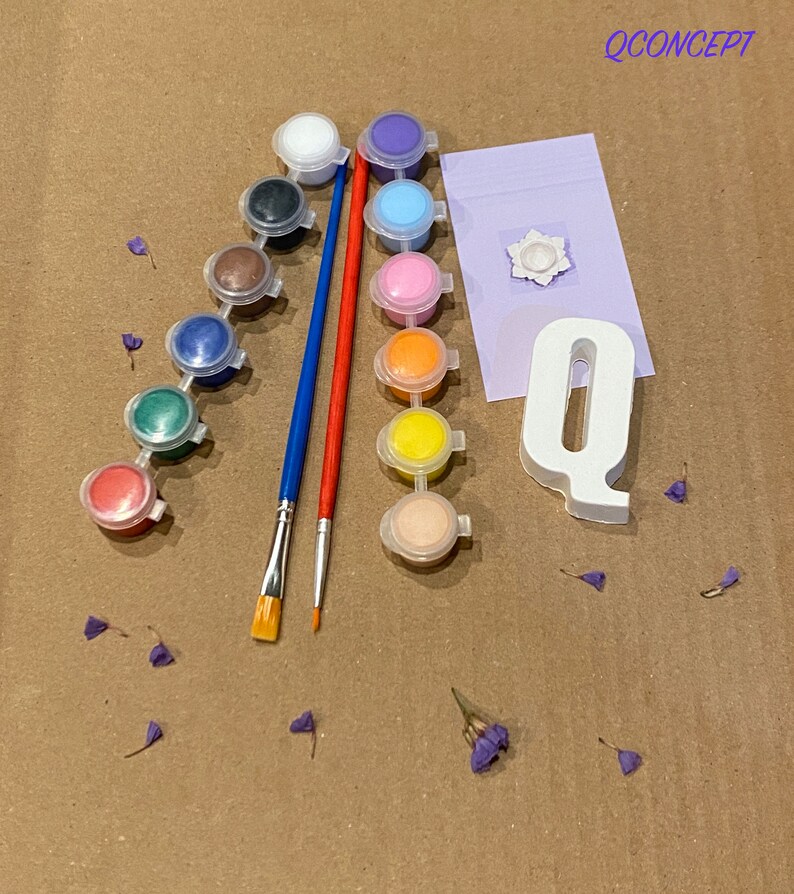 May include: A paint-by-numbers kit featuring a white letter Q, a small white flower, and 12 paint pots in various colours. The kit also includes two paintbrushes, one red and one blue.