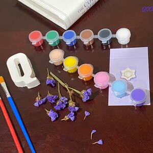 May include: A set of paint pots with various colours, including red, green, blue, yellow, orange, pink, and purple. There are two paintbrushes, one red and one blue. A white ceramic letter Q, a small white flower, and purple flowers are also included.