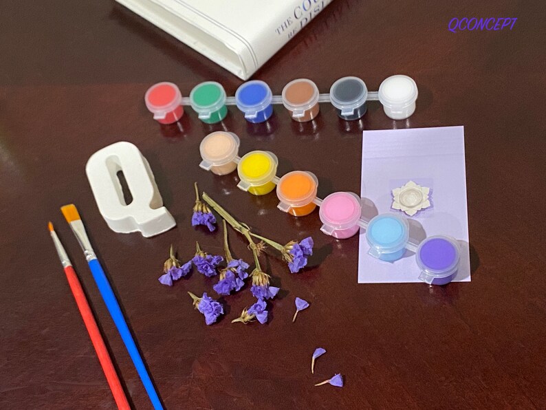 May include: A set of paint pots with various colours, including red, green, blue, yellow, orange, pink, and purple. There are two paintbrushes, one red and one blue. A white ceramic letter Q, a small white flower, and purple flowers are also included.