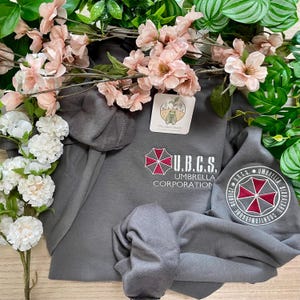 May include: A gray sweatshirt with the Umbrella Corporation logo and the text "U.B.C.S." in white. The sweatshirt has a round patch on the sleeve with the Umbrella Corporation logo and the text "Umbrella Biohazard Countermeasure Service".