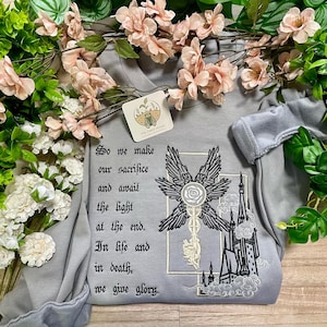 May include: A light gray sweatshirt with embroidered design and text. The design features a key with wings, a rose, and a castle. The text reads "So we make our sacrifice and await the light at the end. In life and in death, we give glory."