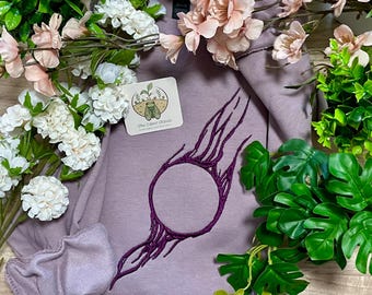 Könnte beinhalten: Ein malvenfarbenes Langarm-Sweatshirt mit einem dunkelvioletten, gestickten kreisförmigen Design mit strahlenförmigen Linien. Ein kleines quadratisches Etikett am Stoff zeigt eine Zikaden-Illustration und den Text The Silent Cicada Embroidery and Illustration.