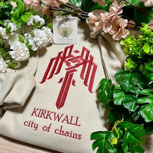 May include: A beige hoodie featuring a large red embroidered design and the words "KIRKWALL city of chains" in red. The hoodie is surrounded by flowers and foliage, with a small tag at the top.