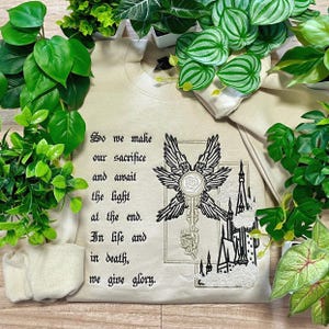 Könnte beinhalten: Beiges Sweatshirt mit gesticktem Design. Das Design zeigt einen Schlüssel mit Engelsflügeln, ein Schloss und den Text "So we make our sacrifice and await the light at the end. In life and in death, we give glory."