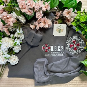 Może przedstawiać: Szara bluza z kapturem z napisem "U.B.C.S. Umbrella Corporation" i czerwono-białym logo parasola. Bluza ma naszywkę na rękawie z napisem "Umbrella Biohazard Countermeasure Service". Różowe i białe kwiaty otaczają bluzę.