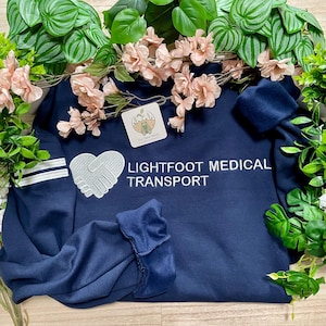 Può includere: Una felpa blu navy a maniche lunghe con un logo ricamato bianco a forma di cuore che raffigura una stretta di mano e il testo LIGHTFOOT MEDICAL TRANSPORT. La manica presenta due strisce bianche orizzontali vicino al polsino.