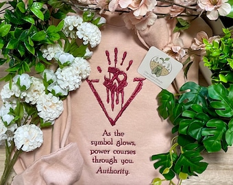 May include: A light pink sweatshirt featuring dark red embroidery of a handprint symbol inside a triangle, with the text: As the symbol glows, power courses through you. Authority. A small square tag with a cicada illustration is attached.