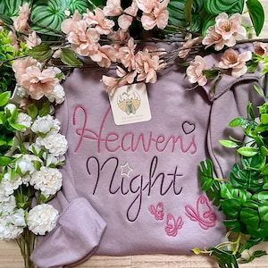 Puede incluir: Una sudadera malva con el texto bordado "Heaven's Night" en rosa oscuro y morado, acompañado de un pequeño corazón, una estrella y tres contornos de mariposas. Una etiqueta cuadrada con una ilustración de una cigarra está unida al cuello.