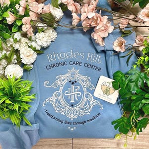 以下が含まれることがあります： ライトブルーのフード付きスウェットシャツ。ネイビーブルーで「Rhodes Hill Chronic Care Center」と刺繍されています。白いエンブレムと「Transforming lives through medicine」の文字があります。小さなタグが付いています。