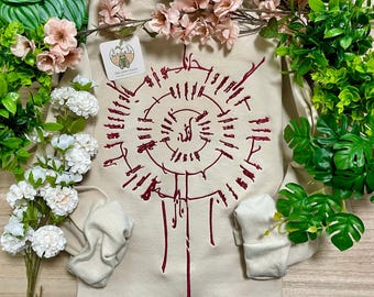 May include: A beige long-sleeve sweatshirt featuring a dark red circular graphic design with intricate, radiating lines and a central symbol. A small square tag with a green insect illustration and the text The Silent Cicada is attached.