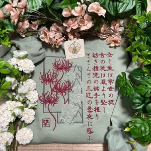 May include: A sage green sweatshirt featuring detailed embroidery of red spider lilies, white cloud outlines, and vertical Japanese text. The design includes a small square red stamp at the bottom left and a small square brand tag at the top centre.