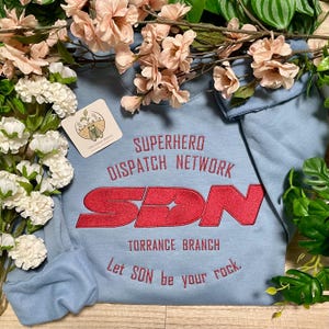 May include: A light blue sweatshirt featuring red embroidered text that reads SUPERHERO DISPATCH NETWORK, SDN, TORRANCE BRANCH, and Let SDN be your rock. A small square tag with a graphic of a cicada and the text The Silent Cicada is attached.