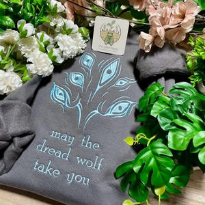 Peut inclure: Un sweat-shirt gris foncé avec une broderie bleu clair représentant une plume de paon et l'inscription "may the dread wolf take you". Le logo The Silent Glade est visible. Le sweat-shirt est entouré de fleurs et de feuillages artificiels.
