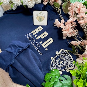Può includere: Felpa blu navy con ricamo "R.P.D. Raccoon City Police Department" e stemma della polizia. La felpa ha girocollo e maniche lunghe. Il design è color crema.