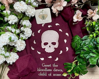May include: A maroon sweatshirt featuring a light pink embroidered skull surrounded by small petal shapes. Below the skull, the text reads: Greet the bloodless dawn, child of none. A small square tag with a cicada logo is attached to the fabric.