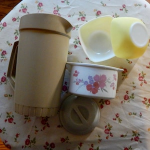 May include: A set of four kitchenware items in beige and white. The set includes a large pitcher, a small bowl with a floral design, a small white bowl, and a lid.