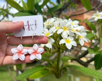 Puede incluir: Un par de pendientes de flores blancas con centros rosas. Los pendientes cuelgan de aros dorados. Los pendientes están delante de una tarjeta blanca con el texto "MJ Designs" en ella. La tarjeta está siendo sostenida por una mano.