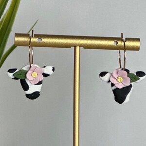 May include: A pair of gold hoop earrings with black and white cow print charms and pink flower accents.