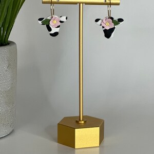 May include: A pair of gold hoop earrings with black and white cow-print charms and pink flowers. The earrings are displayed on a gold stand.