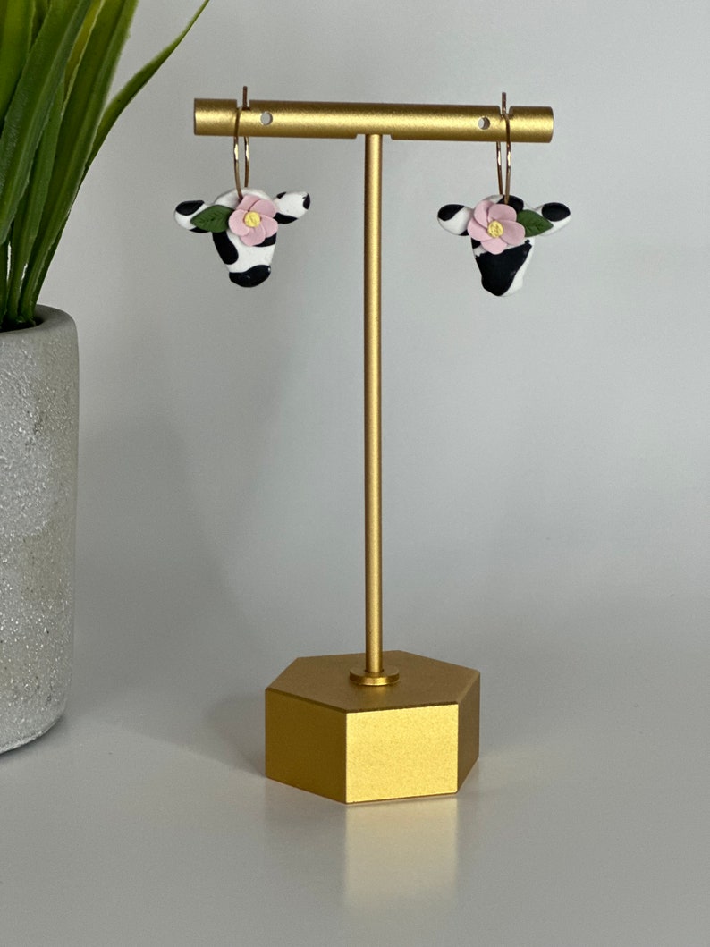 May include: A pair of gold hoop earrings with black and white cow-print charms and pink flowers. The earrings are displayed on a gold stand.