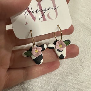 May include: A pair of gold hoop earrings with black and white cow print charms and pink flowers. The earrings are on a white card with the text "MJ Designs".