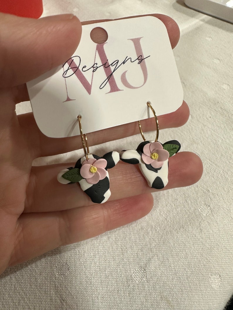 May include: A pair of gold hoop earrings with black and white cow print charms and pink flowers. The earrings are on a white card with the text "MJ Designs".
