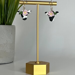 May include: A pair of black and white cow-shaped earrings with pink flowers and green leaves. The earrings are hanging from a gold stand.