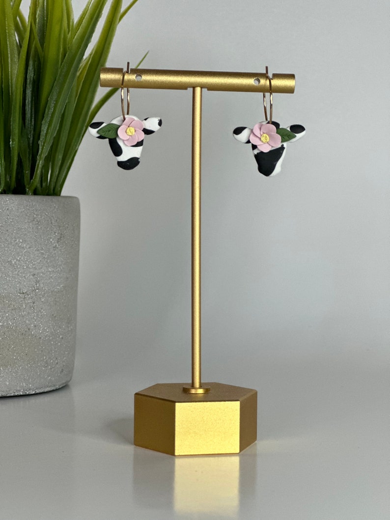 May include: A pair of black and white cow-shaped earrings with pink flowers and green leaves. The earrings are hanging from a gold stand.