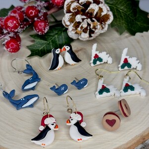 May include: A collection of handmade earrings featuring various designs. Includes whale, penguin, and holly-themed earrings, along with a pair of heart-shaped cookie earrings. The earrings are displayed on a wooden surface with holiday decorations.