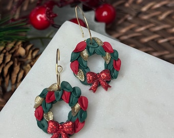 May include: A pair of gold hoop earrings with a red and green Christmas wreath charm. The wreath is decorated with gold glitter and a red bow.