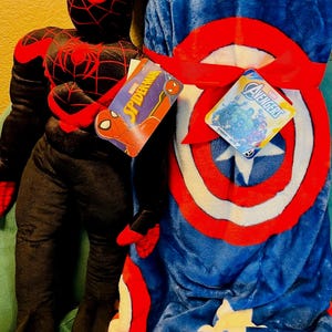 May include: A plush Spider-Man figure in a black and red suit stands next to a blue blanket with a Captain America shield design. The blanket is tied with a red ribbon and has a white star. Tags read "Spider-Man" and "Avengers".