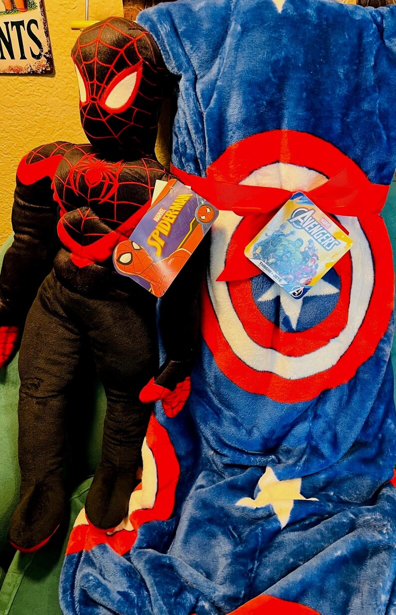 May include: A plush Spider-Man figure in a black and red suit stands next to a blue blanket with a Captain America shield design. The blanket is tied with a red ribbon and has a white star. Tags read "Spider-Man" and "Avengers".