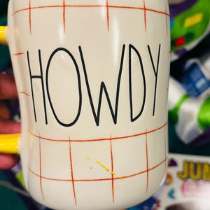 May include: White ceramic mug with the word "HOWDY" in black lettering. The mug has a yellow handle and a red grid pattern. A few yellow paint splatters are visible on the side of the mug.