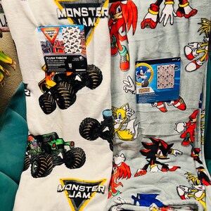 Op de afbeelding: Twee pluche dekens. E&eacute;n is wit met Monster Jam monstertruck graphics en een bijpassende doos. De andere is lichtgrijs met Sonic the Hedgehog karakters en een bijpassende doos. Beide zijn opgevouwen.