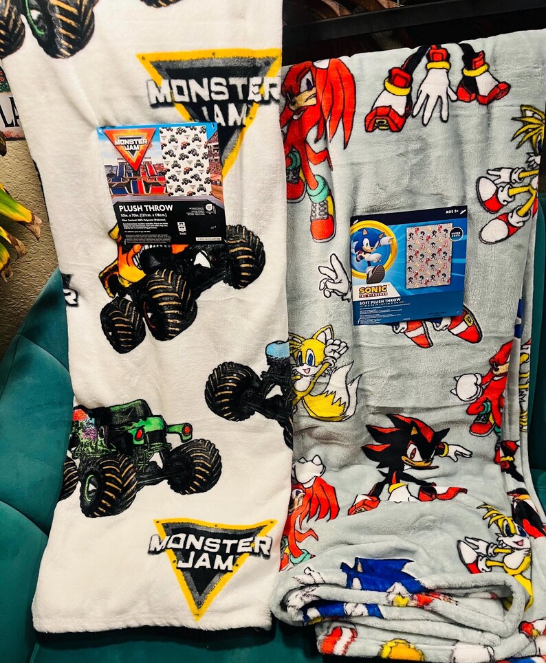 Op de afbeelding: Twee pluche dekens. E&eacute;n is wit met Monster Jam monstertruck graphics en een bijpassende doos. De andere is lichtgrijs met Sonic the Hedgehog karakters en een bijpassende doos. Beide zijn opgevouwen.