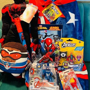 May include: A collection of Marvel Avengers merchandise, including a plush Spider-Man, a Captain America pillow, a blue and red blanket, action figures, a coloring book, and a lollipop. The items are arranged on a teal surface.