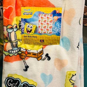 May include: A soft plush throw blanket featuring SpongeBob SquarePants characters. The blanket has a white background with orange, peach, and blue heart shapes. The packaging shows the blanket's design and the words "Soft Plush Throw".
