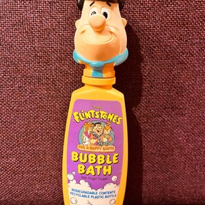 May include: A vintage Flintstones bubble bath bottle with a Fred Flintstone finger puppet topper. The yellow bottle has a purple label with cartoon characters and the text "BUBBLE BATH". The bottle contains 8 fl oz (237 ml) of bubble gum scented bubble bath.