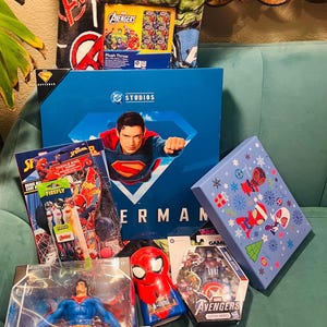 Peut inclure: Divers produits d&eacute;riv&eacute;s sur le th&egrave;me des super-h&eacute;ros, dont un coffret Superman, une couverture Avengers et des jouets Spider-Man. Les articles sont pr&eacute;sent&eacute;s sur une surface turquoise, avec des couleurs et des motifs vari&eacute;s.