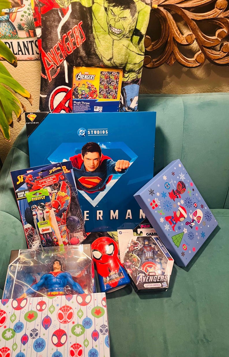 Peut inclure: Divers produits d&eacute;riv&eacute;s sur le th&egrave;me des super-h&eacute;ros, dont un coffret Superman, une couverture Avengers et des jouets Spider-Man. Les articles sont pr&eacute;sent&eacute;s sur une surface turquoise, avec des couleurs et des motifs vari&eacute;s.
