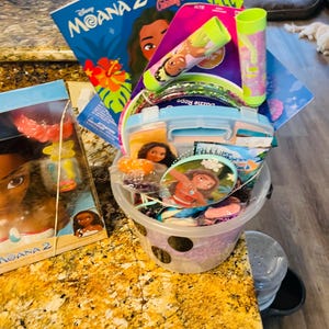 Puede incluir: Una colecci&oacute;n de art&iacute;culos con tem&aacute;tica de Moana, que incluye un libro para colorear, pulseras, una botella de agua y una lonchera. Los art&iacute;culos est&aacute;n en un cubo transparente con dise&ntilde;o de lunares y una lonchera azul. El libro para colorear se titula "Moana 2 Jumbo Coloring and Activity Book."