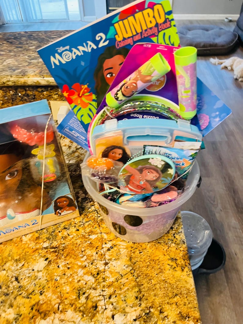 Puede incluir: Una colecci&oacute;n de art&iacute;culos con tem&aacute;tica de Moana, que incluye un libro para colorear, pulseras, una botella de agua y una lonchera. Los art&iacute;culos est&aacute;n en un cubo transparente con dise&ntilde;o de lunares y una lonchera azul. El libro para colorear se titula "Moana 2 Jumbo Coloring and Activity Book."