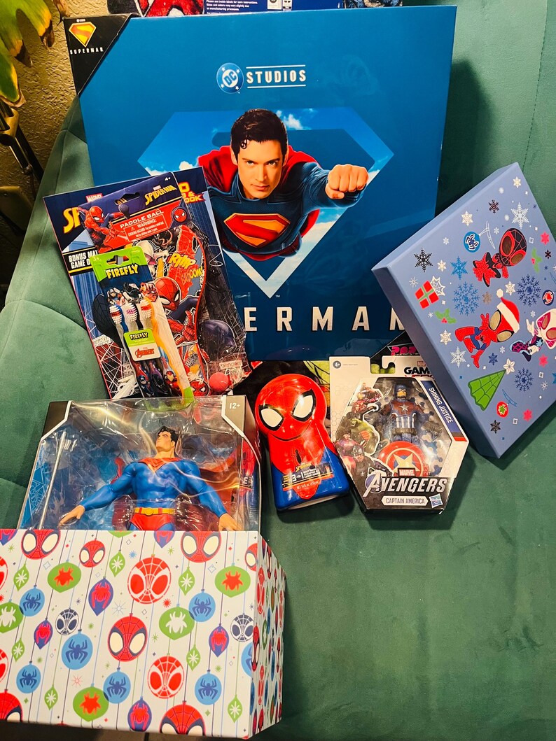 Peut inclure: Plusieurs articles sur le th&egrave;me des super-h&eacute;ros, dont un coffret Superman, des figurines et des jouets Spider-Man. Les articles sont dans des bo&icirc;tes et des emballages vari&eacute;s, avec des couleurs vives et des graphismes de personnages. Le texte "DC Studios" et "Avengers" est visible.