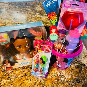 May include: A collection of Disney Moana-themed items, including a doll, coloring book, and a bucket filled with treats. The bucket features a Moana graphic and contains a water bottle, lollipops, and a light-up toy. A Pop Ups! lollipop is also visible.