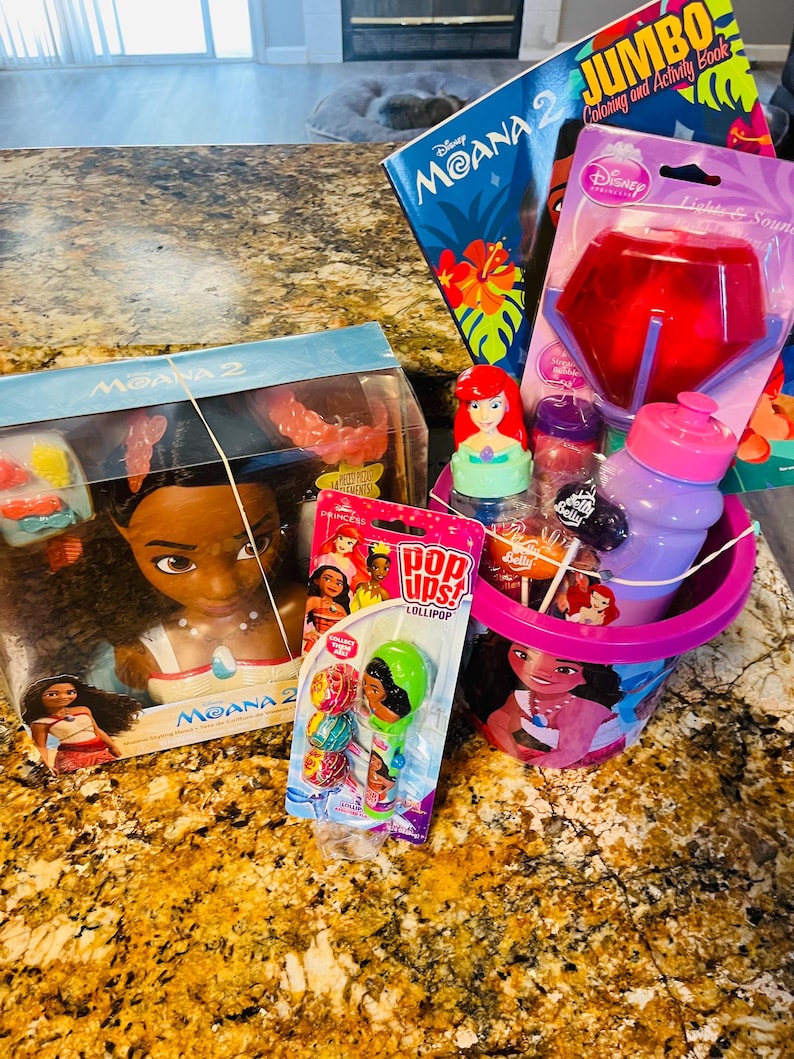 May include: A collection of Disney Moana-themed items, including a doll, coloring book, and a bucket filled with treats. The bucket features a Moana graphic and contains a water bottle, lollipops, and a light-up toy. A Pop Ups! lollipop is also visible.